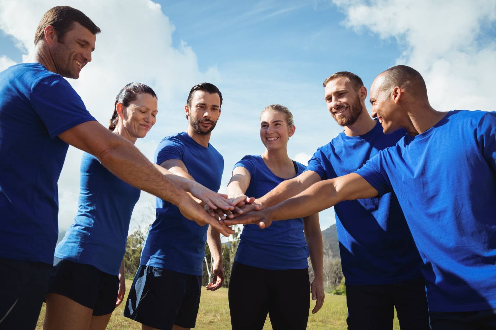 Cinco adultos de camisetas azuis juntam as mãos em um gesto de equipe ao ar livre.